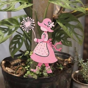 Peut inclure: Une décoration de jardin rose en forme de personnage portant un bonnet et une robe à pois, tenant une tête de pissenlit. Le personnage est représenté de profil avec une expression faciale simple, des bottes roses et une écharpe flottante.