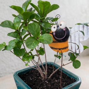 Peut inclure: Une décoration de jardin en métal en forme d'abeille avec un corps rayé noir et orange, un visage souriant aux grands yeux blancs et des ailes blanches texturées. L'abeille est placée parmi les feuilles vertes d'une plante en pot.