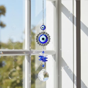 May include: A decorative hanging suncatcher with an evil eye motif. The suncatcher features a blue and white eye design, set within a silver-toned metal frame. Below the eye are blue crystal elements and a clear crystal ball.