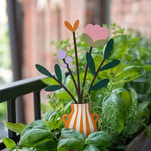 Può includere: Un tutore decorativo in metallo per piante con una base a forma di vaso a righe arancioni e bianche, sormontato da un bouquet di elementi floreali piatti e colorati, tra cui un fiore rosa, un piccolo cuore arancione e bacche viola.