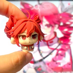 Puede incluir: Una cabeza de figura en miniatura de estilo anime, con cabello y ojos rojos. La figura se sostiene entre los dedos, mostrando sus detalles. El fondo es una imagen borrosa de un personaje similar.