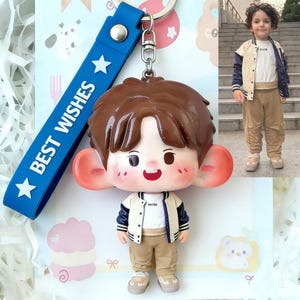 May include: A keychain featuring a cartoon boy with large ears, wearing a white jacket with navy blue sleeves and khaki pants. The keychain has a blue strap with the words "BEST WISHES" and white stars. The boy's hair is brown, and he has a smiling face.