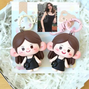 May include: Two keychains featuring a cartoon girl with brown hair, large ears, and a black dress. Each keychain has a matching color ring. The background includes a photo of a woman in a black dress.