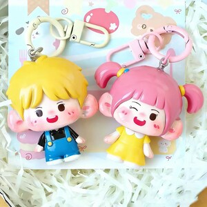 May include: Two cartoon character keychains. One keychain features a boy with yellow hair wearing blue overalls. The other keychain features a girl with pink pigtails wearing a yellow dress. Both have large ears and are smiling.