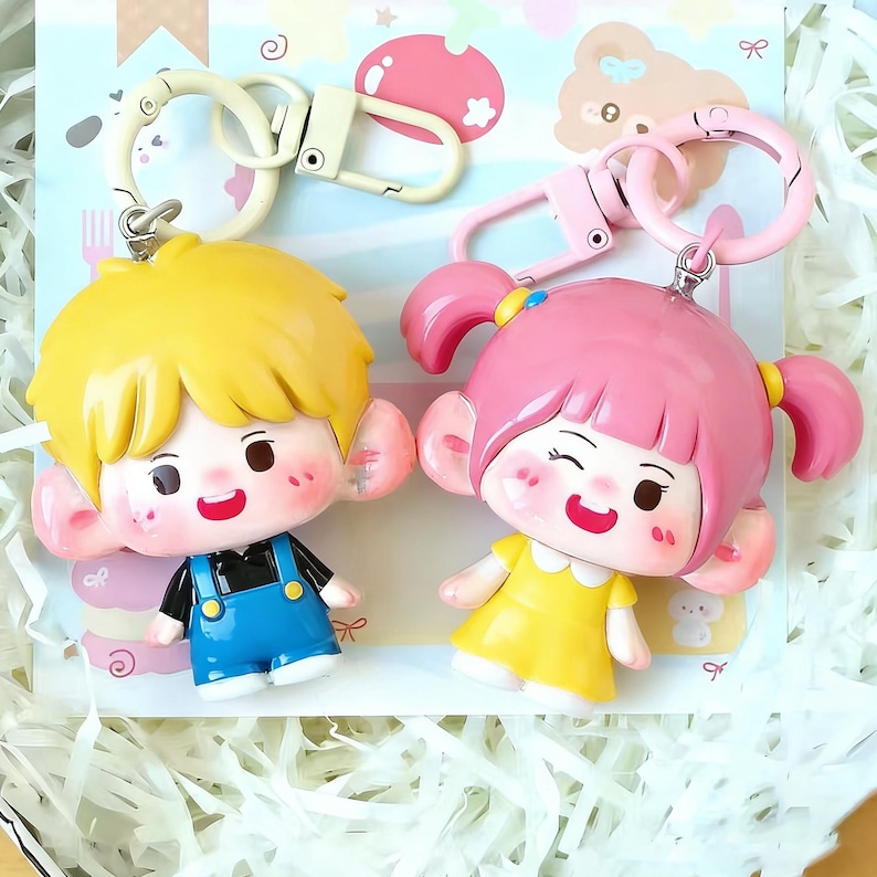 May include: Two cartoon character keychains. One keychain features a boy with yellow hair wearing blue overalls. The other keychain features a girl with pink pigtails wearing a yellow dress. Both have large ears and are smiling.
