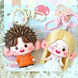 May include: Two cartoon character keychains. One keychain features a boy with brown hair and an orange hoodie. The other keychain features a girl with blonde hair and a white dress. Both keychains have large ears and are attached to metal clasps.