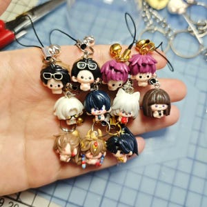 Puede incluir: Una colección de pequeños amuletos de personajes de estilo anime coloridos. Cada amuleto presenta un peinado y una expresión facial únicos, unidos a un cordón negro con una campana plateada o dorada. Los amuletos se sostienen en una mano.