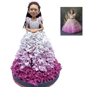 May include: A cake topper featuring a young girl in a white and pink ombre dress. The dress is made of layered fabric, and the girl has long dark hair and a tiara. A smaller image of the girl in the dress is in the background.