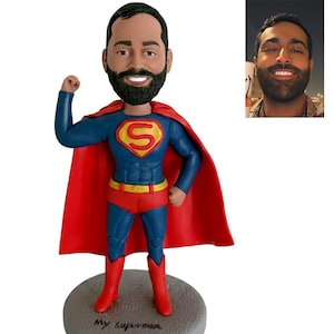 May include: A Superman bobblehead figurine with a custom face. The figure wears a blue suit with a red cape, boots, and briefs. The chest features a yellow and red "S" emblem. The base is grey and has the words "My superman" written on it.