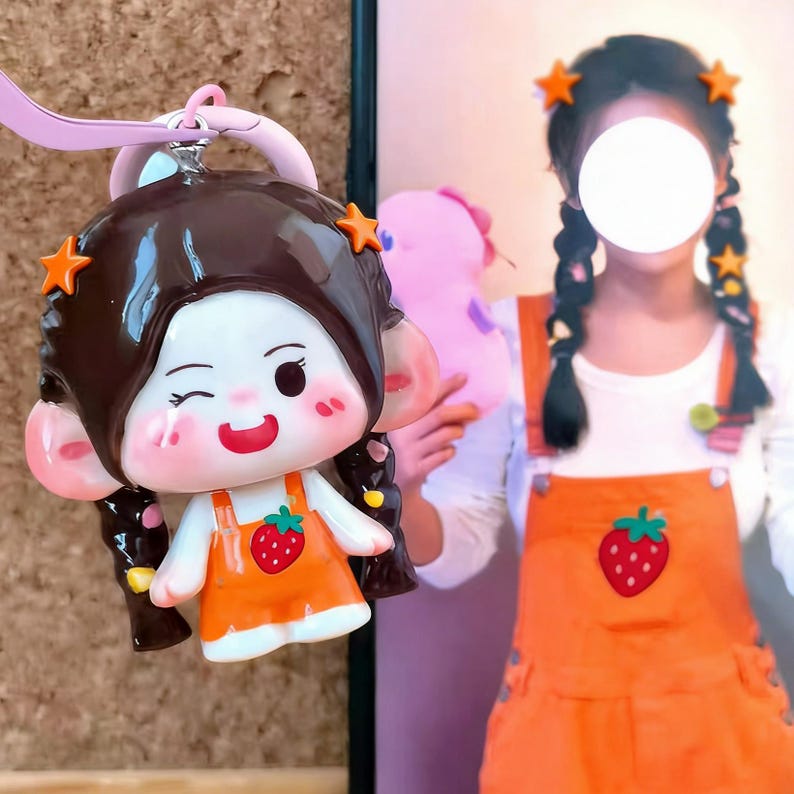 May include: A charming keychain featuring a cartoon girl with brown hair, orange star hair clips, and an orange dress with a strawberry design. The keychain has a pink strap and a silver metal ring. The girl is winking and smiling.