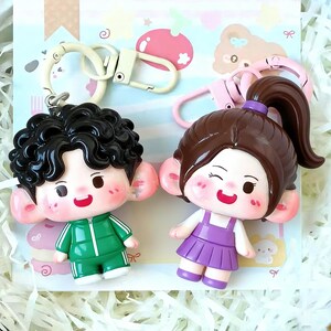 May include: Two keychains featuring cartoon figures. One figure has black curly hair and wears a green tracksuit. The other figure has brown hair in a ponytail and wears a purple dress. Both have rosy cheeks and large ears. Each keychain has a metal clasp.