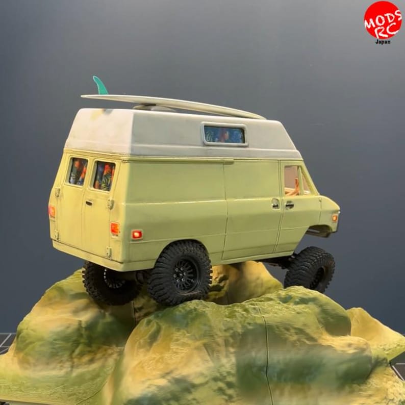 May include: A pale yellow scale model van with a white high-top roof and a teal surfboard mounted on top. The vehicle features black off-road tyres, orange taillights, and a side window. The text MODS RC Japan is visible in the top right corner.