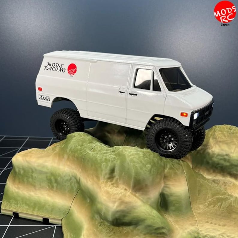 May include: A white remote-controlled van model with black off-road tires and wheels. The side features the text Mods Racing and a red circular logo. The van has orange side marker lights and a black front grille. It is positioned on a textured terrain.