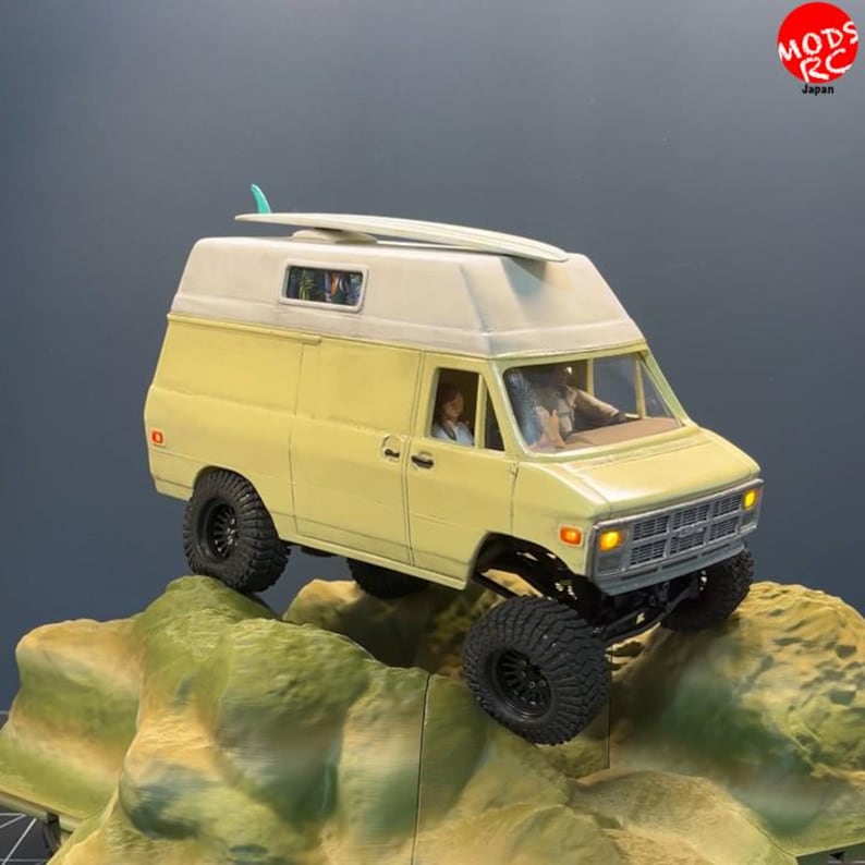 May include: A yellow and white scale model camper van with a surfboard on the roof, featuring black off-road tyres, a silver grille, and illuminated amber headlights. The vehicle is positioned on a textured, rocky terrain base with a driver visible.