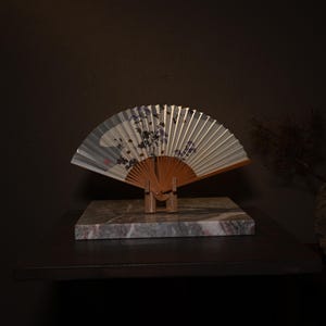May include: A decorative hand fan with a floral design in shades of purple and gray, displayed on a marble block. The fan is made of paper and wood, and is open to reveal its full design. The fan is propped up by a wooden stand.