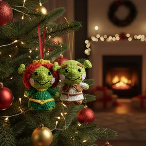 Pode incluir: Dois enfeites de Natal feitos à mão em crochê em forma de ogro verde e sua companheira, pendurados em uma árvore de Natal. Os enfeites têm detalhes em vermelho, verde e amarelo. A árvore é decorada com bolas vermelhas e douradas e luzes.