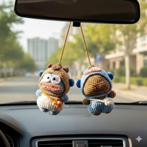 May include: Two handmade crochet car charms hanging from a rearview mirror. One charm is brown and blue, the other is blue and white. Both charms are in the shape of a cartoon character with large eyes and a small bag.