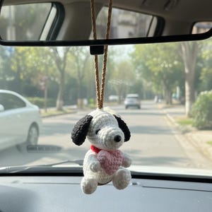 Puede incluir: Un adorno de coche de perro de ganchillo blanco y negro con un corazón rosa y un collar rojo, colgado del espejo retrovisor de un coche. El adorno está suspendido por una cuerda marrón.