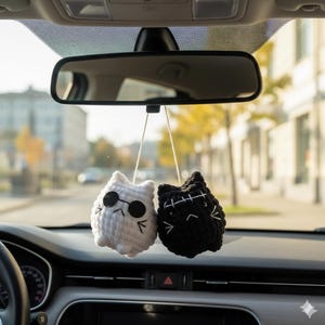 Puede incluir: Dos amuletos de coche de gato de ganchillo colgando del espejo retrovisor de un coche. Un gato es blanco con gafas de sol negras, el otro es negro con costuras blancas. Se ve el salpicadero y el parabrisas del coche.
