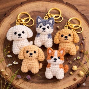 May include: Five crochet dog keychains in various colors: white, gray, and orange. Each keychain features a gold-colored ring. The dogs have black eyes and noses, and are arranged on a wooden surface.