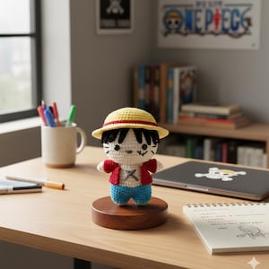 May include: A crocheted Luffy doll from the One Piece anime series. The doll has a yellow hat with a red band, a red jacket, blue shorts, and black hair. It is displayed on a wooden stand on a wooden desk. A mug with pens and a laptop are in the background.
