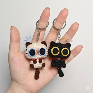 以下が含まれることがあります： 2つの手編みの猫のキーホルダー。1つはクリーム色で茶色のアクセントと青い目、もう1つは黒で黄色の目です。どちらも赤い首輪とシルバーのキーリングが付いています。キーホルダーは手作りで、漫画の猫のデザインが特徴です。