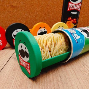 Może przedstawiać: Zielona puszka Pringles z tacą chipsów wystającą na zewnątrz. Kilka kolorowych pokrywek Pringles znajduje się w tle. Widoczne jest również logo Pringles i czarna puszka Pringles.