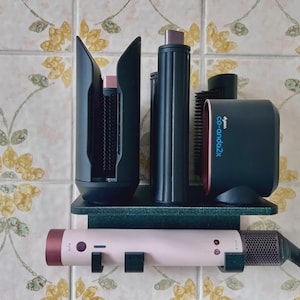 May include: A black wall-mounted storage rack holding three black hair styling attachments and a Dyson Coanda 2x smoothing dryer. A light pink hair styling wand with a dark red end cap and a black power cord rests horizontally on the rack's base.
