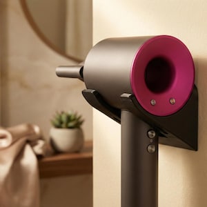 May include: A dark gray hair dryer with a magenta accent, mounted on a black wall bracket. The hair dryer is cylindrical with a nozzle and button controls. The bracket is attached to a beige wall.