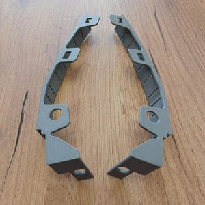 May include: Two identical gray, L-shaped brackets with a textured finish. Each bracket features a long, curved arm with multiple rectangular cutouts along the inner edge and two circular mounting holes positioned on the lower section of the frame.