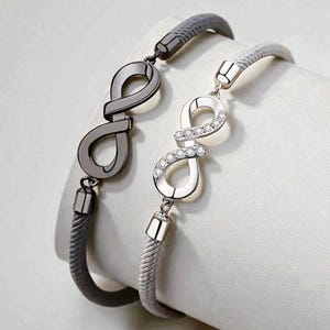 Könnte beinhalten: Zwei Armbänder mit Unendlichkeitszeichen. Ein Armband hat ein dunkelgraues Unendlichkeitszeichen mit passendem grauem Kordel. Das andere Armband hat ein silbernes Unendlichkeitszeichen mit klaren Edelsteinen und einem hellgrauen Kordel. Beide Armbänder haben silberne Verschlüsse.