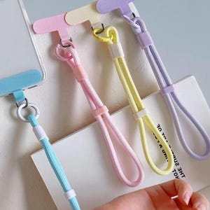 May include: A clear smartphone case with a light blue adhesive tab attached to a light blue wrist lanyard. Three additional wrist lanyards in pastel pink, yellow, and lavender are displayed, each featuring a matching T-shaped adhesive tab.