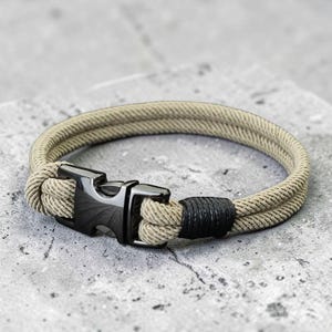 Puede incluir: Una pulsera de cuerda beige con un cierre de metal negro. La pulsera est&aacute; hecha de dos hebras paralelas de cuerda, aseguradas con un cierre negro y una banda negra. La pulsera tiene una circunferencia de aproximadamente 20 cm.