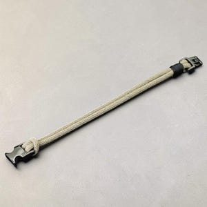 Puede incluir: Una pulsera de paracord beige y negro con un cierre negro. La pulsera est&aacute; hecha de dos cordones paralelos, anudados en un extremo y asegurados con un cierre de hebilla negro en el otro. La pulsera mide aproximadamente 20 cm de largo.