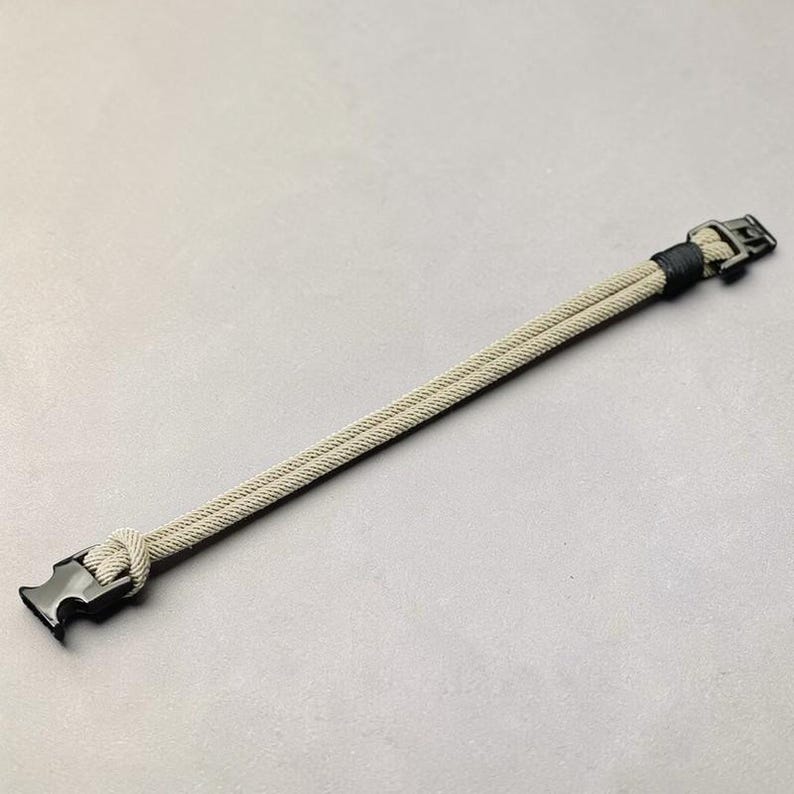 Puede incluir: Una pulsera de paracord beige y negro con un cierre negro. La pulsera est&aacute; hecha de dos cordones paralelos, anudados en un extremo y asegurados con un cierre de hebilla negro en el otro. La pulsera mide aproximadamente 20 cm de largo.