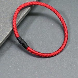 May include: A red braided bracelet with a black clasp. The bracelet is circular and appears to be made of a woven material. The clasp is a simple, rectangular design. The bracelet is on a grey surface.