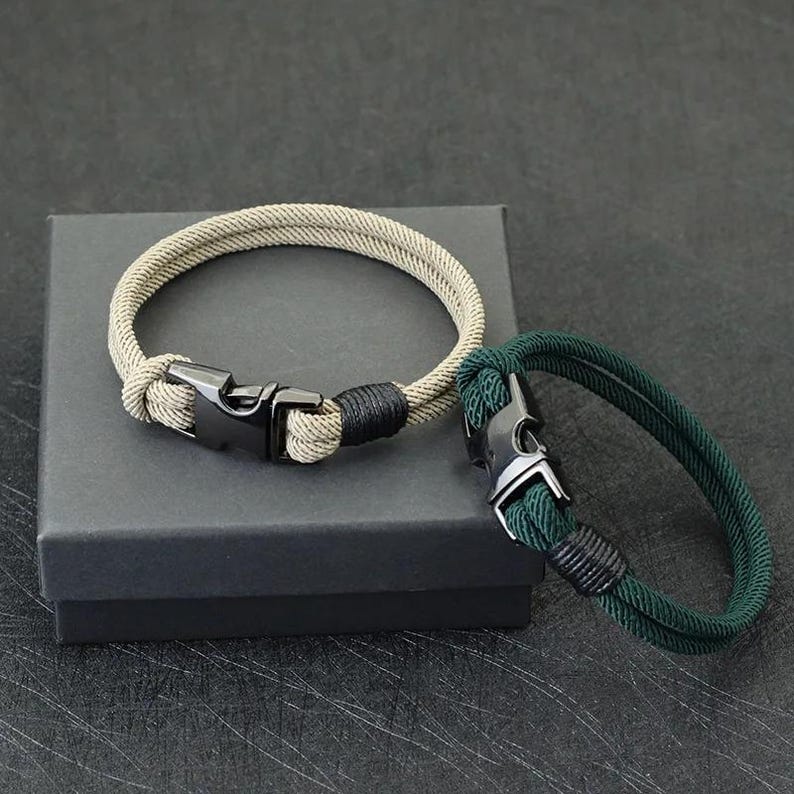 Puede incluir: Dos pulseras de cuerda. Una es de color beige con un cierre y detalles en negro, la otra es verde oscuro con un cierre plateado y detalles en negro. Ambas pulseras est&aacute;n sobre una caja gris oscura.