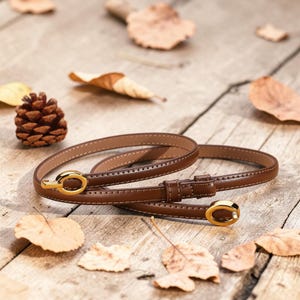 May include: A thin brown leather belt featuring a polished gold-tone oval buckle and a matching gold-tone loop. The belt has visible stitching along the edges and a smooth finish, designed with a slim profile for a refined and clean appearance.