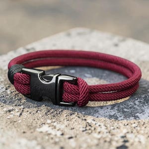 Puede incluir: Una pulsera de cuerda burdeos con un cierre de metal negro. La pulsera est&aacute; hecha de dos hebras paralelas de cuerda burdeos, aseguradas con una hebilla de metal negro. La pulsera se muestra sobre una superficie gris texturizada.