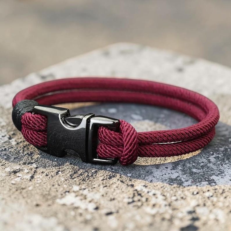 Puede incluir: Una pulsera de cuerda burdeos con un cierre de metal negro. La pulsera est&aacute; hecha de dos hebras paralelas de cuerda burdeos, aseguradas con una hebilla de metal negro. La pulsera se muestra sobre una superficie gris texturizada.