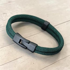 May include: A dark green double-strand cord bracelet featuring a matte black plastic side-release buckle clasp. The bracelet consists of two parallel woven cords joined by a matching black plastic sliding keeper loop for a secure and simple design.