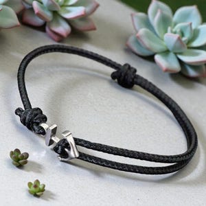 Puede incluir: Pulsera de cuerda negra con cierre de ancla plateado. La pulsera está anudada en ambos extremos y presenta un diseño de doble hebra. El cierre de ancla es un detalle prominente. El fondo incluye plantas suculentas.