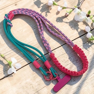 May include: A lanyard crafted from teal and multi-colored patterned paracord, featuring a section of pink braided cord and a pink plastic clip attachment. The design includes two small pink metal rings connecting the cords to the clip.