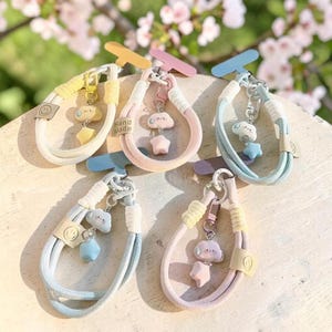 May include: Five phone wrist straps in pastel colors, each featuring a looped cord, a small metal clip, and a charm set consisting of a cloud and a star. Each strap includes a small rectangular tag with a smiley face icon or the text Hand Made.