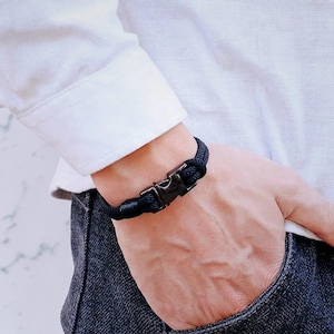 Puede incluir: Pulsera de cuerda negra con cierre met&aacute;lico. La pulsera est&aacute; hecha de una gruesa cuerda negra tejida y presenta un cierre met&aacute;lico seguro. La pulsera se lleva en la mu&ntilde;eca y el cierre est&aacute; abrochado.
