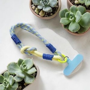 May include: A phone lanyard featuring a light blue and yellow patterned rope with solid blue cord wrappings and a bright yellow knot. A light blue T-shaped plastic attachment is connected to the end of the rope by a small metal ring.