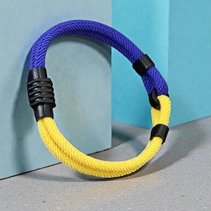 May include: A blue and yellow rope bracelet with black connectors. The bracelet is made of a thick, woven rope material. The bracelet is a vibrant accessory.