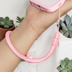 May include: A light pink braided wrist strap for a mobile phone, featuring a matching pink cylindrical connector and a small sliding adjustment bead. The strap is constructed from a woven cord material and attaches to a pink phone case.