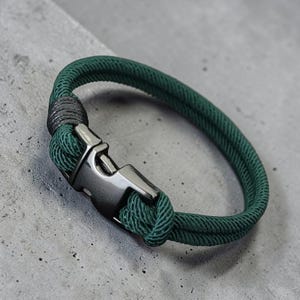 Puede incluir: Una pulsera de cuerda verde con un cierre de metal de ca&ntilde;&oacute;n de fusil. La pulsera est&aacute; hecha de dos hebras de cuerda verde, anudadas en un extremo. El cierre tiene forma rectangular con un cierre seguro.