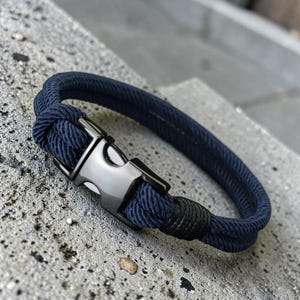 Puede incluir: Pulsera de cuerda azul marino con cierre met&aacute;lico plateado. La pulsera est&aacute; hecha de un material de cuerda gruesa y tejida. El cierre tiene forma rectangular con un cierre seguro. La pulsera se muestra sobre una superficie de hormig&oacute;n gris.